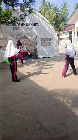 This video captures an outdoor scene on a bright, sunny day, likely in a tropical or warm climate, as indicated by the presence of palm trees. The ground is made of dirt or sand, and long shadows suggest midday.

The central focus is a large, white canvas tent, which appears to be a temporary structure. Printed prominently on the tent are the words "KEMENDIKDASMEN" (likely an abbreviation for the Ministry of Education and Culture in Indonesia), along with the phrase "PENDIDIKAN BERMUTU UNTUK SEMUA" (Quality Education for All) and "RAMAH" (Friendly/Welcoming). There is also a logo featuring a cross and a heart, possibly indicating a health or humanitarian initiative. Inside the tent, stacks of chairs or stools are visible, and an adult woman stands in the doorway.

Several children, primarily girls, are present. They are dressed in school uniforms, consisting of black pants and white hijabs, with some wearing pink or red accents on their clothing. In the foreground, two girls are engaged in what appears to be a game; one holds a long stick, while the other adopts a ready stance. Another child is seen running in the background near a building that could be part of a school. The children seem to be playing cheerfully in the outdoor environment.