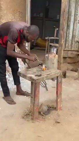 A medium shot shows a man working outdoors with a blowtorch. He wears a red and black striped athletic shirt with "ETIHAD AIRWAYS" on the front, black sweatpants, and sandals. He has a beard and a wide smile. He works at a small, square metal table, and the torch he holds emits a large, bright flame. Behind him, another man walks into a building. The ground is dirt. A skinny wooden post stands to the right of the table.