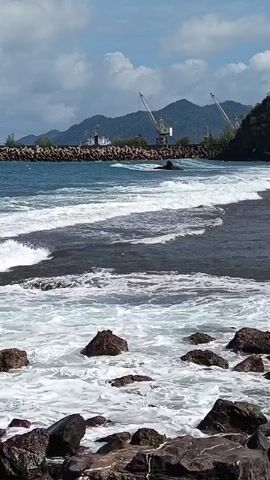The video displays a coastal scene with a body of water in the foreground. Waves are breaking against a rocky shoreline, with white foam covering the rocks and water. In the middle ground, there is a breakwater made of large stones, with the sea on the other side. Beyond the breakwater, a ship is visible, and further in the distance, there are two large cranes. Mountains form the backdrop under a partly cloudy sky. The video appears to be taken from a stationary viewpoint, possibly handheld, and the focus is on the ocean and its interaction with the shore and man-made structures. There are no people visible in the video. The overall impression is a dynamic seascape with a hint of industrial activity in the background.