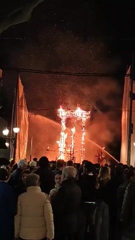 At night, a large crowd watches as a tall, wooden structure burns in the middle of a street. Two streams of water are being sprayed on the fire. The people in the crowd are of various ages and are wearing dark clothing. They are standing close together and looking towards the fire. There are streetlights and tall, white banners on either side of the street. Small embers are flying in the air.