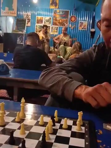 This is a first-person perspective video showing a chess board in the foreground, with a lively scene in the background. Two men are playing acoustic guitars and singing, while other patrons appear to be watching them. The setting seems to be an indoor cafe or bar, possibly in Bhagsu, India, given the mention of the location context. The walls are decorated with colorful artwork and posters, some depicting fantastical or mythological scenes. The lighting suggests it could be evening or late afternoon. The overall atmosphere appears relaxed and convivial. The chess board is set up with pieces in the middle of a game, with white appearing to have the advantage. The camera person's hand is visible on the right side of the frame, suggesting they might be playing chess or are a spectator. There is some visible text on a poster on the back wall which reads "COWLE WORLD".