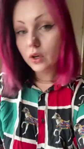 A person with bright pink hair and a shirt with a multicolored pattern that includes horses looks directly at the camera. She flips her hair back. The background is unclear.