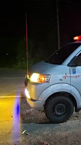 A white ambulance with red and blue flashing lights is parked on a dirt road at night. The ambulance has a heartbeat graphic on its side. The front of the ambulance is illuminated by its headlights, and there are also yellow and blue lights on the ground in front of it. A person's hand is visible reaching out of the driver's side window. The background is dark and shows trees and a pole.