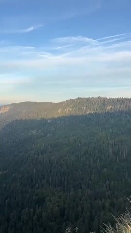 The video displays a panoramic aerial view of a lush green mountainous landscape under a clear blue sky with scattered clouds. The terrain is characterized by steep, forested slopes and deep valleys. As the camera pans, a prominent, rocky cliff face becomes visible on the left side of the frame. The light suggests either sunrise or sunset, casting long shadows across the mountains and highlighting the contours of the landscape. In the distance, a hazy valley with hints of human settlement is discernible. The video consists of four distinct segments, each capturing a slightly different angle or portion of the expansive natural scenery.
