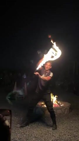 A man dressed in a dark t-shirt and pants is performing a fire-spinning act at night. He is in a circular area with a campfire in the center. He is holding two long objects that are on fire and spinning them around himself and in front of him, creating streaks of light. There are people sitting in chairs in the background, watching the performance. The ground is covered in gravel.