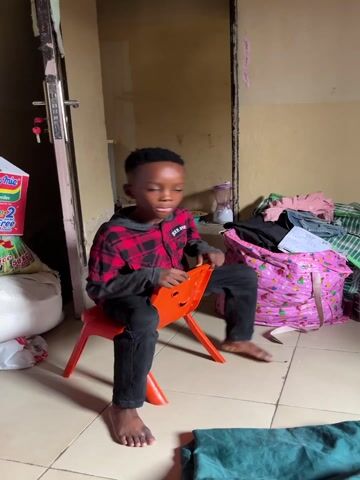 A young boy, barefoot, with short dark hair, is seen indoors in what appears to be a home in Gauraka, Nigeria. He is wearing a red and black plaid shirt over a dark grey long-sleeve shirt (with "JOKER SPIRIT" visible on the chest) and dark pants. He sits on an orange plastic child's chair, which has a smiling face design on its back. Throughout the video, the boy rocks back and forth on the chair, holding onto the top of its back.

The room has light-colored walls, a tiled floor, and appears somewhat cluttered. Various items are visible in the background: a large pink bag with white patterns filled with clothes, a green and white striped bag, a pile of fabrics, and a woven mat or fabric with a yellow and brown pattern. On the left, near a door with a silver handle and a key, a white bag with "CE" and "NGS" visible on it is present. A blender, a plastic jug, and a red plastic bag are also seen in the background. A green blanket or piece of fabric lies on the floor in the foreground. The lighting suggests daytime. The boy looks towards the camera briefly at the end of the video.