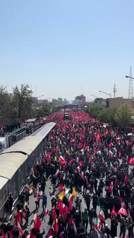 A large crowd of people are walking down a street, holding many red flags. Some of the flags are green, white, and red, which appears to be the flag of Iran. The crowd is dense, filling the entire width of the street, and extends far into the distance. There are buildings and trees visible on either side of the street, and a clear blue sky above. In the distance, there is a truck with people standing on it. The scene appears to be a procession or a large gathering.