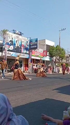 A street parade is taking place with several people in elaborate costumes. The costumes appear to be traditional or ceremonial, featuring rich fabrics, intricate patterns, and headdresses. One individual in the foreground is holding a framed portrait of a man. There are buildings on the side of the street with signage, suggesting a commercial area. Spectators are seated on the side of the road, some with drinks and snacks. The sky is clear and blue, indicating daytime. Motorcycles are parked along the street. The overall scene suggests a cultural event or festival.