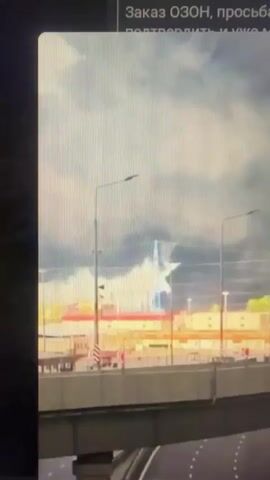The video shows a view from a moving vehicle, looking towards a cityscape under a cloudy sky. In the foreground, the edge of a road and a barrier are visible. Further ahead, there is an elevated structure, possibly an overpass or bridge, with streetlights. Beyond that, buildings and industrial structures are visible. A prominent feature is a tall, cylindrical structure with red and white stripes, likely a smokestack or tower, emitting a plume of dark smoke that expands into the sky. The smoke appears to be originating from behind or around the industrial buildings. The overall scene suggests an industrial area experiencing some form of event causing smoke emission. The lighting indicates it is daytime.