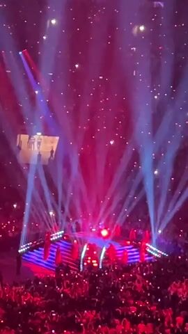 The video shows a concert with a stage lit by red and blue spotlights. A crowd of people, illuminated by phone lights, is visible in the foreground. On the stage, there are performers on risers, and a large screen displays silhouetted figures. The overall atmosphere is energetic and vibrant, with beams of light cutting through the darkness.