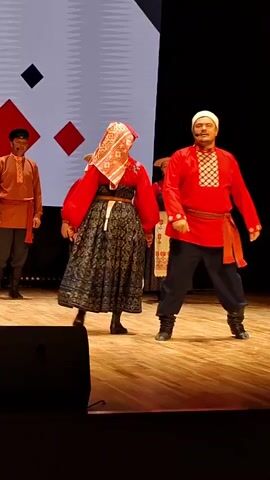 A group of five people are performing on a wooden stage in front of a dark background and a large screen with geometric patterns. The three individuals in the foreground are dressed in traditional folk costumes. On the left, a man in a reddish-orange tunic with a black cap and dark pants stands facing the center. In the center, a man in a bright red tunic with intricate white embroidery on the collar and chest, black pants, and a white cap is facing a woman. The woman is wearing a red blouse, a white apron with embroidered patterns, a dark patterned skirt, and a headscarf. The man and woman are holding hands and appear to be in the middle of a dance or performance. To the right of the couple, another woman in a similar folk costume is visible, also in red and with a headscarf. The overall atmosphere appears to be a cultural performance or theatrical event.