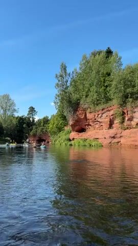 The video shows a wide river with calm water flowing from left to right. On the right bank, there are red rock formations covered in green vegetation and trees. Several people are in kayaks on the river, appearing to be on a leisurely outing. The sky is clear and blue. The overall scene is natural and peaceful.