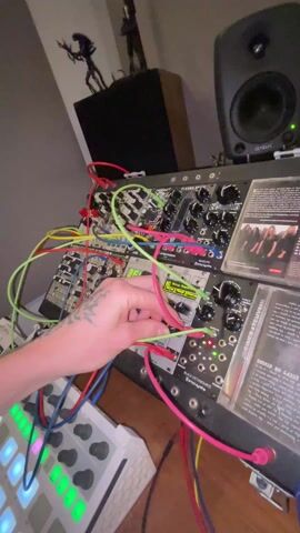 The video shows a person operating a modular synthesizer. The synth, placed on a wooden surface, is cluttered with colorful patch cables. The person's left hand is visible, adorned with a tattoo on the wrist and forearm. They are seen adjusting the green patch cables on the synth, specifically around the module labeled "Black Octasource by Erica Synths". A white MIDI controller sits beside the synth, with various lit buttons in different colors. In the background, a black speaker is visible, with a green light indicating it is powered on. Behind the speaker, there are figurines placed on a high shelf. Next to the synthesizer are two CD cases. The overall environment looks like a home music studio setup.
