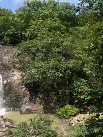 The video displays a waterfall cascading down rocky cliffs surrounded by lush green trees under a partly cloudy sky. Several people are visible in the pool at the base of the waterfall, some swimming and others standing on the rocks. The water is a murky green color, and the rocks are dark and wet from the spray. The scene appears to be a natural outdoor environment, possibly a park or wilderness area, and the activity suggests recreational use of the waterfall.