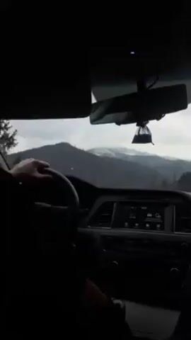 The video shows the interior of a car driving on a road. A hand is visible on the steering wheel. The dashboard and central console are in clear view. The car's infotainment screen is on. Outside the car's front windshield are mountains, some with snow on top, visible in the distance under a cloudy sky.