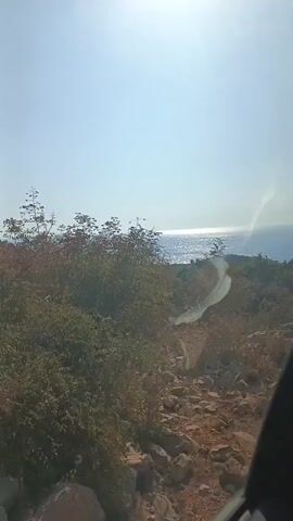 The video shows a view from a moving vehicle looking out at a rocky, sparsely vegetated hillside with the ocean in the distance under a bright sun. The camera is angled slightly downwards and to the right. The sun is high in the sky, creating a glare on the lens. The ocean is visible as a wide expanse of blue water with sunlight glinting off its surface. There are scattered bushes and rocks on the hillside in the foreground and middle ground. The video appears to be shot from inside a vehicle, with a portion of the car's interior visible at the bottom right of the frame. The overall impression is one of a scenic drive along a coastal area.