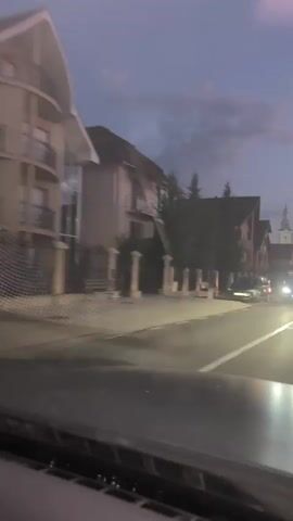 A car is driving down a street at dusk. The street is wet, reflecting the headlights of oncoming vehicles and streetlights. There are buildings on either side of the street, some with multi-story facades. A church steeple is visible in the distance. The sky is overcast and the light is dim. The video is shot from inside the car, looking out through the windshield.