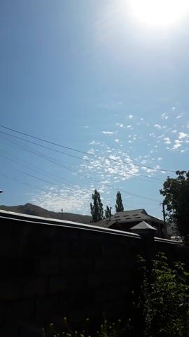 This media file depicts an outdoor scene on a bright, sunny day in Gul'cha, Kyrgyzstan. The image is oriented vertically. The sky is a clear, vibrant blue with scattered, wispy white clouds. The sun is a prominent, radiant disc on the left side of the frame, casting a lens flare and strong rays, suggesting it is daytime, likely mid-morning or afternoon given the brightness.

In the background, a dark, silhouetted structure, possibly a building with multiple windows and a pitched roof, is visible. Several trees with lush green foliage are present, with some partially obscuring the building. Power lines stretch horizontally across the sky, adding a sense of human infrastructure to the natural landscape. A mountain range, also in silhouette, can be seen in the far distance, beneath the clouds and behind the power lines.

The foreground is dominated by a dark, textured surface, which appears to be a wall or a fence, casting a shadow and framing the lower right portion of the image. Some yellow flowers are visible at the very bottom right, adding a small touch of color. The overall impression is one of a peaceful, rural or semi-rural environment bathed in sunlight. There is no visible text in the media file.