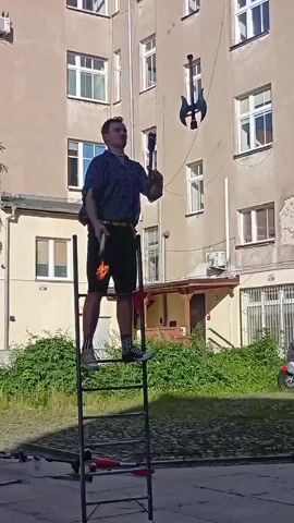 A young man is standing on a ladder and juggling two axes and one club. The man is wearing a blue dotted shirt, black shorts, and colorful socks. He is standing on a metal ladder with red grips. Behind him is a beige building with multiple windows. The ground in front of the ladder is covered with a black mat, and there is green grass and bushes to the right of the mat. There is also a scooter in the background to the right. The man is in the process of juggling the axes and club, with one axe in his left hand, one axe in the air, and the club in the air.