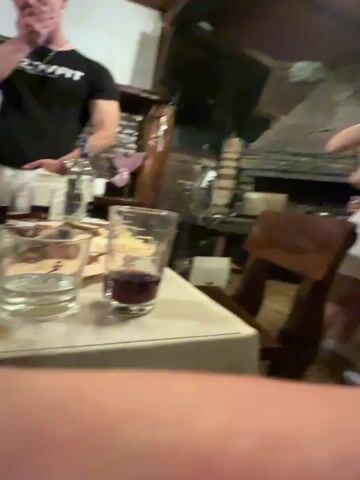The video shows an indoor scene with several people. A man in a black t-shirt and white pants is standing and appears to be talking or thinking. Another man in a white shirt is visible in the background. On a table in the foreground, there are glasses, some containing liquid, plates, and utensils. The environment appears to be a restaurant or bar, with some furniture and decorations visible in the background. The camera is close to the table, and the focus is on the objects on the table and the man in the black t-shirt.