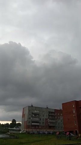 The video captures a view of a cloudy sky with large, dark clouds overhead. In the foreground, there are two apartment buildings. The building on the left is light gray with some reddish-brown accents and several stories. The building on the right is made of red brick and also has multiple stories. Below the buildings, there is a grassy area with some parked cars visible. The overall impression is of an overcast day, possibly before or during a storm. There are no people or animals clearly visible, and no explicit actions are taking place.