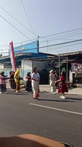 A group of women and girls are walking in a procession down a street. Many of the participants are playing musical instruments, including drums. They are dressed in traditional clothing. A large Indonesian flag is being carried by one of the women. The procession is moving from left to right in the frame. The background shows a blue building with a sign, as well as other structures and utility poles. The sky is clear and the sun is shining. The camera appears to be mounted on a vehicle, as indicated by the slight vibration and the bottom edge of what looks like a seat or dashboard.