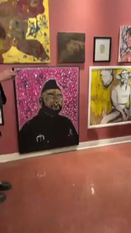 The video shows an interior view of an art gallery with red walls and a red-patterned floor. The walls are adorned with various paintings and framed artwork. The camera pans slowly across the wall, highlighting several pieces including a large, vibrant painting with gold and abstract shapes, a portrait of a man in a black outfit with a pink background adorned with sparkles, and a framed image of a skeletal figure. The overall impression is that of a contemporary art exhibit.