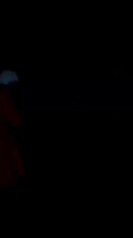 The video shows a group of people in a dimly lit environment. There are indistinct figures, some wearing light-colored clothing, moving about. The overall visibility is low.