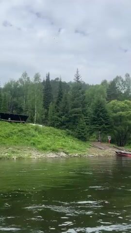 The video begins with a wide shot of a calm, green river with trees and a cloudy sky in the background. The camera then pans to show the shore, with a grassy embankment and more trees. As the video progresses, the perspective shifts to a first-person view from a paddleboard. Another paddleboard is visible to the left, with a person's legs and a bag on it. The camera is focused on the front of the paddleboard, showing a paddle resting on it. The scene appears to be a serene natural environment, possibly a river flowing through a forest. The overall atmosphere is peaceful and relaxed, suggesting a leisurely activity like paddleboarding.