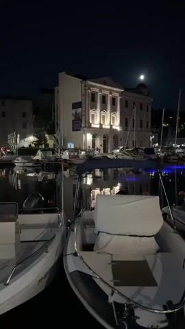 The video shows a nighttime view of a harbor with several boats docked. In the background, a building with neoclassical architecture is illuminated by artificial lights. A full moon is visible in the dark sky above. The water reflects the lights from the building and the boats. The camera pans slightly to the right, showing more of the docked boats and a glimpse of more buildings along the waterfront. The overall atmosphere is calm and serene.