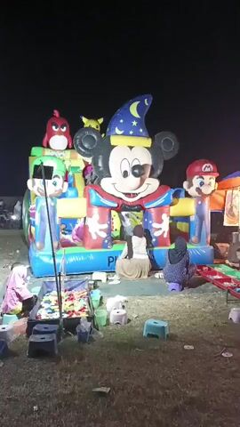 The video shows an inflatable play structure shaped like Mickey Mouse in a wizard hat, with characters resembling Mario and Luigi on either side. Several children are visible playing inside the structure. There are also two women sitting on the ground in front of the play structure. A table with small plastic balls is also present. The setting appears to be an outdoor event at night, as indicated by the darkness and artificial lighting.