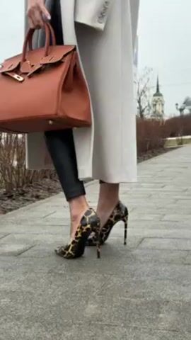 The video shows a woman standing outside on a grey brick walkway in a park or city square. She is wearing black leather leggings, leopard print high heels, a white shirt or top, and a light grey overcoat. She is holding a brown handbag with gold hardware. The woman is adjusting sunglasses on her face. In the background, there are trees, a church or monument with a spire, and an overcast sky. The woman is the central focus and the camera moves slightly to follow her motion.