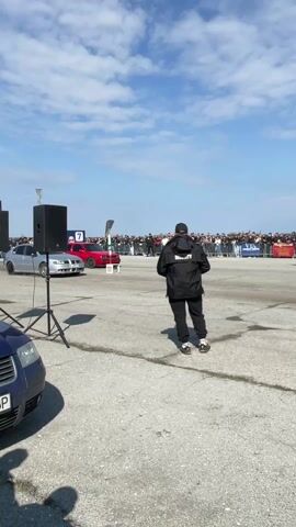 The video shows a drag race between a red hatchback and a silver sedan. The cars take off, spinning their tires and kicking up clouds of dust. A crowd of people stands behind a barrier, watching the race, with some recording it on their phones and tablets. A security guard stands in the foreground, facing the race. The sky is blue, and the race occurs on a large concrete surface next to a field.