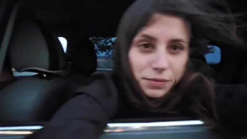 A woman is inside a vehicle, leaning out of the open window. She has long, dark hair and is wearing a dark jacket. Her face is partially obscured by her hair in some frames, and she appears to be making playful expressions, including winking and puckering her lips. The background is blurred, suggesting movement. The lighting is dim, possibly indicating it is dusk or nighttime. The video is shot from a low angle, looking up at the woman.