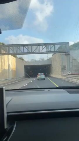 The video shows the view from inside a car as it approaches and enters a tunnel. The car is following another silver car. The tunnel has a concrete ceiling with lights. The walls of the tunnel are light-colored. There is an overhead bridge structure visible before the tunnel entrance.