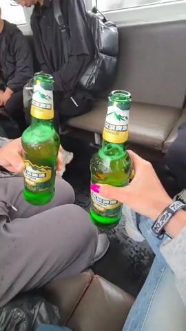 The video captures scenes inside a passenger transport vehicle, likely a ferry, with several people onboard. The setting appears to be during the daytime, possibly under overcast or rainy weather conditions, indicated by the misty view through the windows.

The primary activity shown is two individuals toasting with green glass bottles of Harbin beer, a Chinese brand. One person's hand, holding a bottle, has bright pink nail polish and wears a patterned wristband.

Other passengers are visible throughout the video. They are seated on benches, mostly dressed in casual attire, including black jackets and light-colored clothing. Many are engaged in passive activities such as looking at their mobile phones or resting. One man is seen appearing to sleep, while another woman holds a red bell pepper.

The interior of the vehicle features rows of utilitarian seating. Above the seats, transparent plastic bags filled with various items, possibly luggage or goods, are stored in overhead racks. A "禁止吸烟" (No Smoking) sign, written in Chinese characters, is visible on an interior wall, providing a clear cultural and geographical cue. The overall atmosphere seems relaxed, indicative of a journey. Given the provided location context of Nizhneleninskoe, Russia, this journey is likely a cross-border ferry trip, possibly to or from China, explaining the presence of Chinese beer and signage.