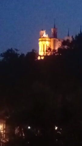 The video shows a dimly lit outdoor scene at dusk or night. A building, possibly a castle or a prominent landmark, is visible on a hilltop in the distance, illuminated by warm yellow lights. The building has multiple towers and ornate architectural features. In the foreground, dark silhouettes of trees and foliage obscure the lower part of the scene. Several small, indistinct lights are scattered in the foreground, suggesting streetlights or other ambient illumination. The sky is a dark blue, indicating twilight. The video appears to be shaky, suggesting it was filmed handheld. No people or specific actions are clearly discernible due to the low light and distance.