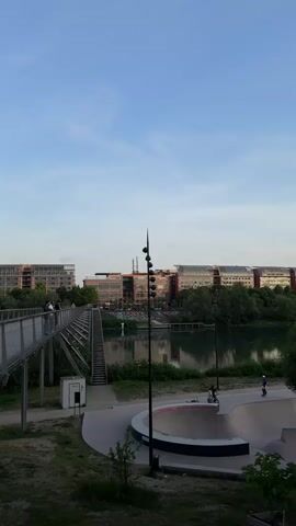 The video captures an expansive outdoor scene in Caluire-et-Cuire, France, under a clear, light blue sky, likely in the late afternoon or early evening. In the foreground on the right, a modern concrete skatepark features a large bowl, with two individuals engaged in activities, possibly skateboarding or cycling. To the left, a contemporary metal pedestrian bridge with stairs extends across a grassy area, with several people walking along its length. A tall, dark, multi-lamp post stands prominently in the center midground. Behind these foreground elements, a calm body of water reflects the sky and distant buildings, with lush green trees and foliage lining its banks. In the background, several large, modern urban buildings with varied architectural styles, including brick and extensive glass facades, line the opposite side of the water. The scene conveys a sense of urban recreation and leisure. No visible text is present.