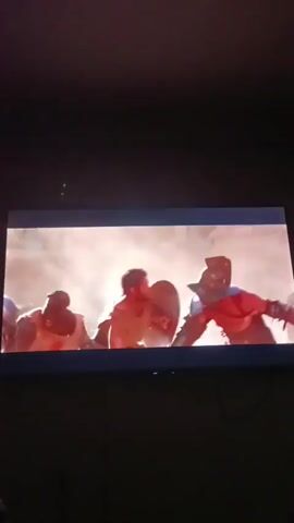 The video shows a scene from a movie or television show, likely a historical drama or action film. There are at least two men in what appears to be an arena or combat setting. One man in the foreground, wearing a tan tunic, is engaged in a fight, raising his arm and holding a shield. He appears to be blocking an attack or defending himself. Another man, partially obscured and wearing armor, is also present and seems to be involved in the combat. The environment is dusty and bright, suggesting an outdoor location. The camera is positioned to capture the action on a screen, likely a television or projector, with a dark room visible around it.