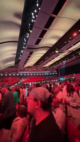 The video shows a large indoor gathering of people, likely a church service or concert, viewed from the perspective of someone in the audience. The ceiling is high and features modern, undulating architectural elements with integrated lighting that casts a red and white glow. Multiple rows of seating are visible, filled with a diverse crowd. On a stage in the distance, there are performers, screens displaying images, and a complex lighting rig with spotlights and colorful effects. The overall atmosphere appears to be one of anticipation and engagement with the event unfolding on stage.