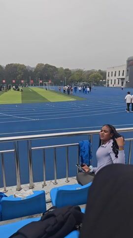 The video shows a view from stadium seating at a track and field event. The track is blue with white lines. A grassy field sits within the track area, with a soccer goal visible. There are people on the track and the field.  A person with braided hair sits in the foreground looking at a phone. Next to them is another person wearing dark pants. The stadium seating is blue. In the distance, there are buildings and trees. The sky is overcast.
