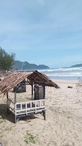 This video showcases a serene beach scene in Meudheun, Indonesia. The primary subjects are the natural elements of the beach: a sandy shore, the vast ocean with rolling waves, and a clear blue sky dotted with clouds. Man-made objects present include a small, rustic wooden hut with a thatched roof, and several picnic tables. The scene is tranquil, with no people visible, suggesting a peaceful and uncrowded environment.

The activity, or lack thereof, points to a moment of quiet observation. The gentle movement of the waves and the slow pan of the camera suggest a relaxed pace. The time of day appears to be daytime, likely morning or early afternoon, given the bright sunlight and the position of the sun, casting shadows. The weather seems pleasant, with a clear sky and visible clouds, indicating a typical tropical day.

Notable details include the distinct hut, which serves as a focal point in the foreground, and the picnic tables that hint at potential visitor activity. The presence of a dry, leafless tree adds a touch of natural artistry to the sandy landscape. The overall mood conveyed is one of peace, natural beauty, and quietude. There is no visible text in the media file. The location context of Meudheun, Indonesia, further grounds the imagery in a specific geographical setting known for its coastal landscapes.