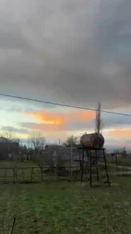 The video shows a cloudy sky with a narrow band of pink and orange color towards the horizon. The foreground features a rusty, horizontal cylindrical tank elevated on a black metal stand in a grassy area. A slender, bare tree stands nearby. There are houses and bare trees visible in the distance. A power line runs across the middle of the frame.