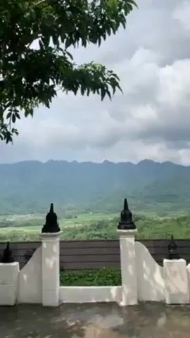 Here is a concise description of the video:

The video shows a scenic view of a valley and distant mountains from behind a decorative white fence. The fence has evenly-spaced posts topped with black, bulbous ornaments. A few tree branches with leaves frame the upper left corner of the shot. The valley is lush and green, and the mountains are hazy with some clouds hanging low. The sky is overcast, suggesting a cloudy day. The foreground surface appears wet, possibly from recent rain.