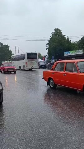 A red sedan is driving on a wet road in front of a white bus. There are other cars and people visible. It appears to be raining.