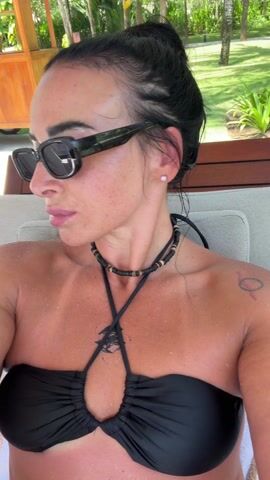 A woman wearing a black strapless bikini top and black sunglasses drinks from a glass of water. Her hair is pulled up in a bun, and she is wearing a beaded necklace. She is sitting on a striped chaise lounge, with a wooden structure and green foliage visible in the background. The woman has freckles on her face and chest. The glass has condensation on the outside. She takes a sip of the water and swallows.