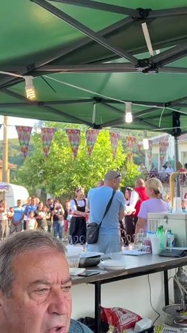 The video shows an outdoor gathering under a green canopy, likely a festival or market. In the foreground, an older man is partially visible, looking down. Behind him, a man in a blue shirt is standing near a table. In the background, a crowd of people are gathered. Some individuals appear to be wearing traditional clothing, including skirts and vests. There are food stalls or trucks visible, with one person actively grilling food. Bunting with balloon designs hangs overhead, and trees and buildings form the backdrop. The lighting suggests it is daytime, possibly late afternoon or early evening.