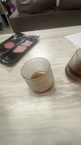 A person is pouring a brown liquid from a clear glass bottle into a decorative glass tumbler containing some of the same liquid. The bottle has a gold-colored ring around its neck. Two such tumblers are on a light-colored, wood-grained table, along with a black plastic tray holding three slices of meat. The background features a brown couch and a person sitting on a chair in the distance.
