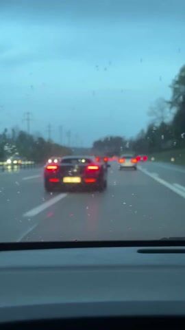 A stationary, close-up shot from the driver's seat shows the rear of a dark-colored McLaren sports car on a highway. The car has its brake lights on and a license plate reading "AG 596 766". It is raining, as evidenced by the droplets on the windshield. Other vehicles are visible in the background. The sky is overcast, suggesting dusk or dawn. Power lines are visible on the side of the road.