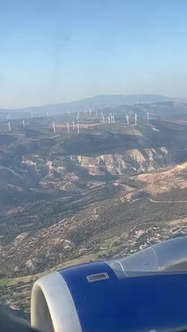 The video shows an aerial view from an airplane window, looking out at a landscape dotted with numerous wind turbines. The turbines are spread across rolling hills and mountainous terrain, some appearing to be on ridgelines. The terrain is a mix of rocky, arid areas with lighter colored soil and vegetated slopes with darker green foliage. In the foreground, a portion of the airplane's engine nacelle, painted blue and silver, is visible. The sky is clear and light blue, suggesting daytime, likely morning or late afternoon due to the angle of the light casting shadows. There are no people or animals clearly visible. The scene appears to be a rural or semi-rural area, potentially dedicated to renewable energy generation.